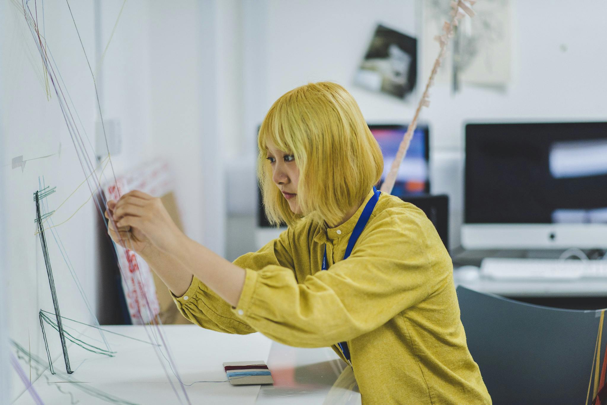 A girl in a yellow top sits at a desk working on an art sculpture built from multicoloured strings.