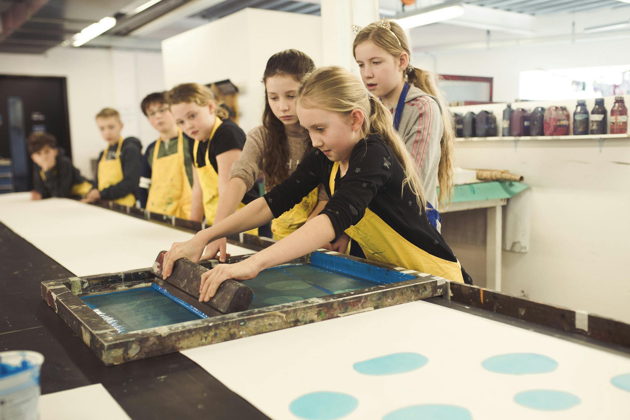 A young girl in a yellow apron holds a squeegee as she pulls it across a printing screen. She's observed by six fellow children in aprons beside a long printing table.