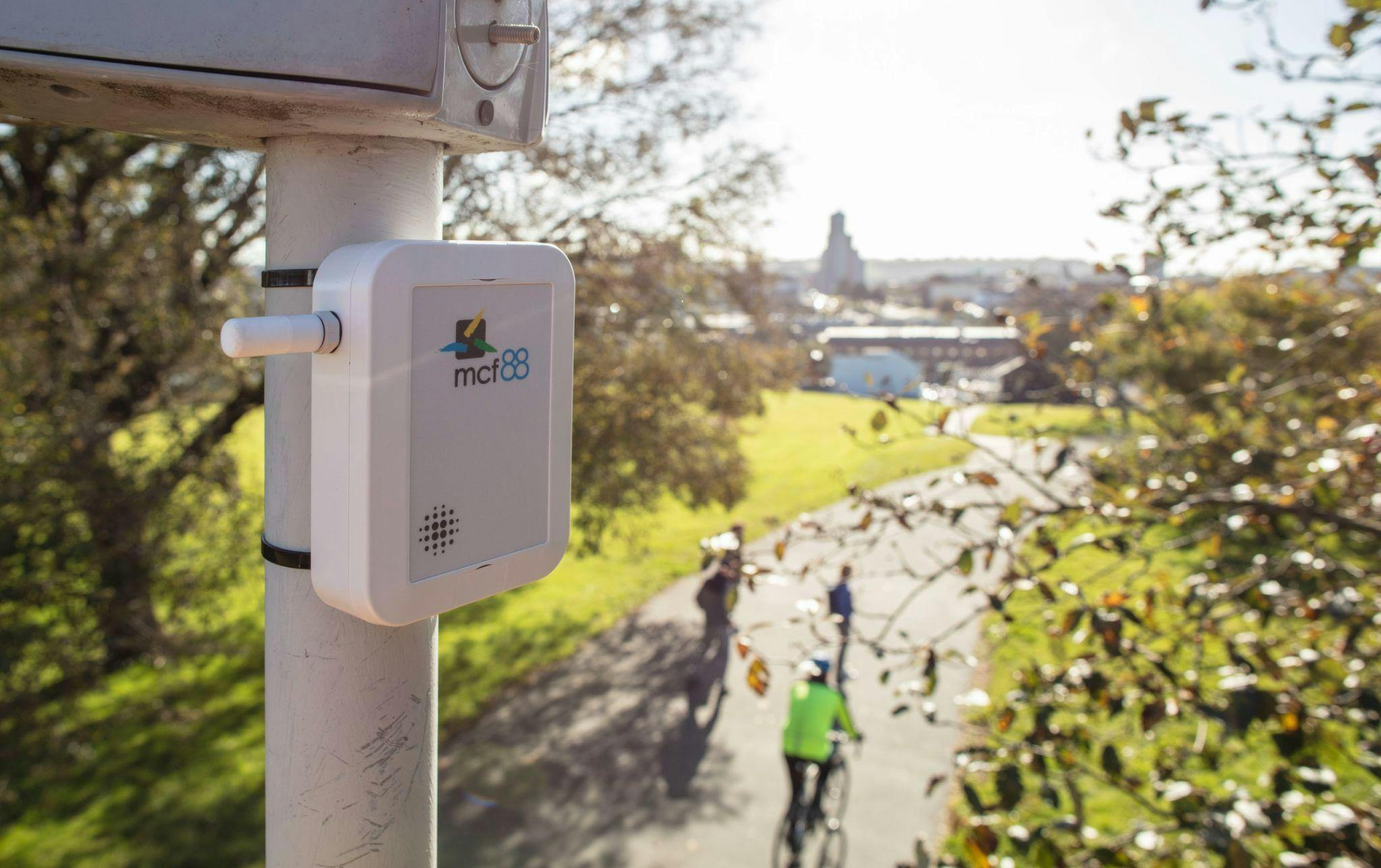 Image of an environmental park sensor high up on a pole overlooking someone riding a bicycle wearing high vis, a couple other people walking and trees and a park in the background