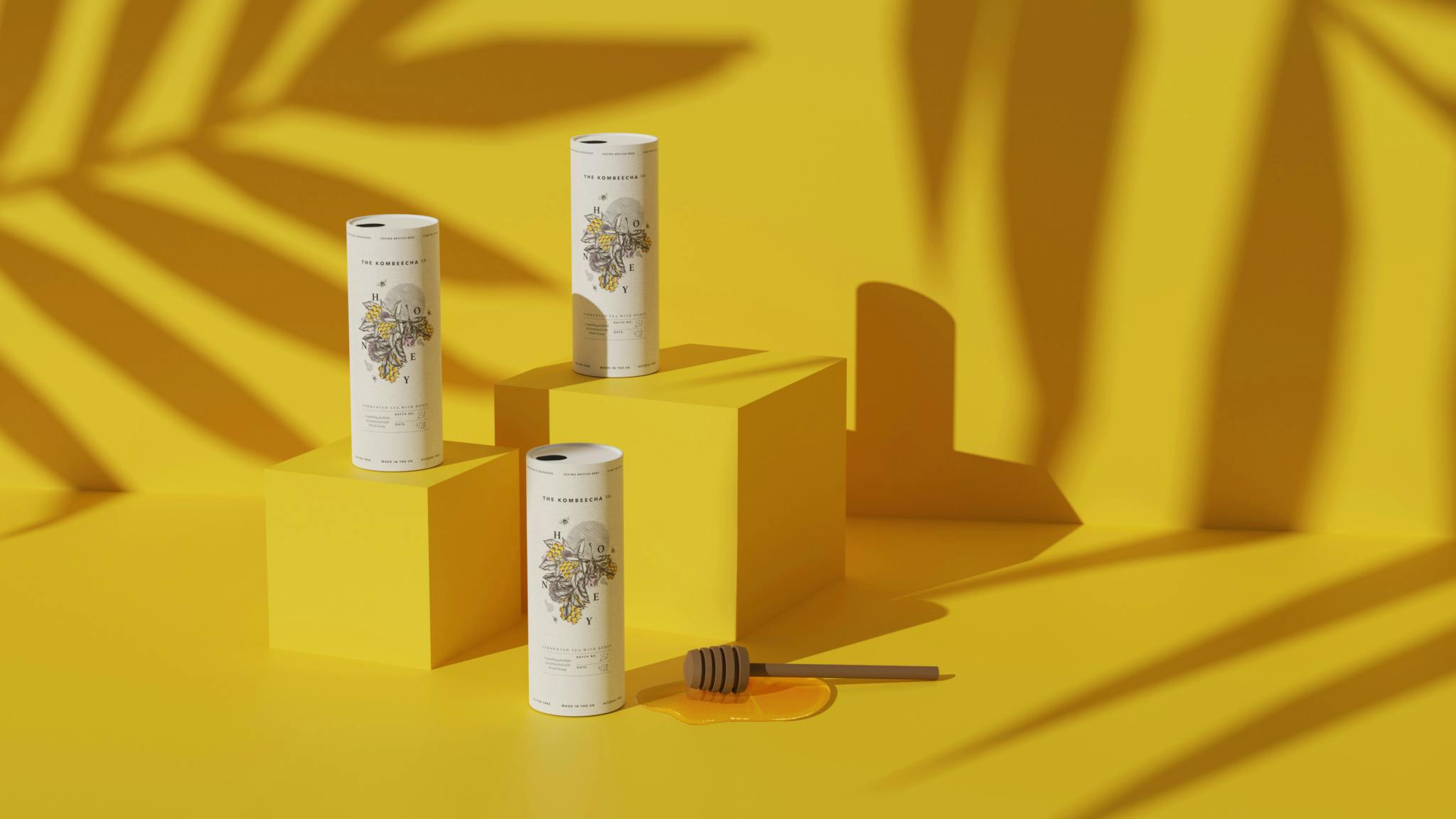 Image shows three kombucha drinks on mustard yellow boxes against a mustard yellow background with shadows of leaves, as well as a honey stirrer in the foreground. The drinks show the illustrated bee branding.