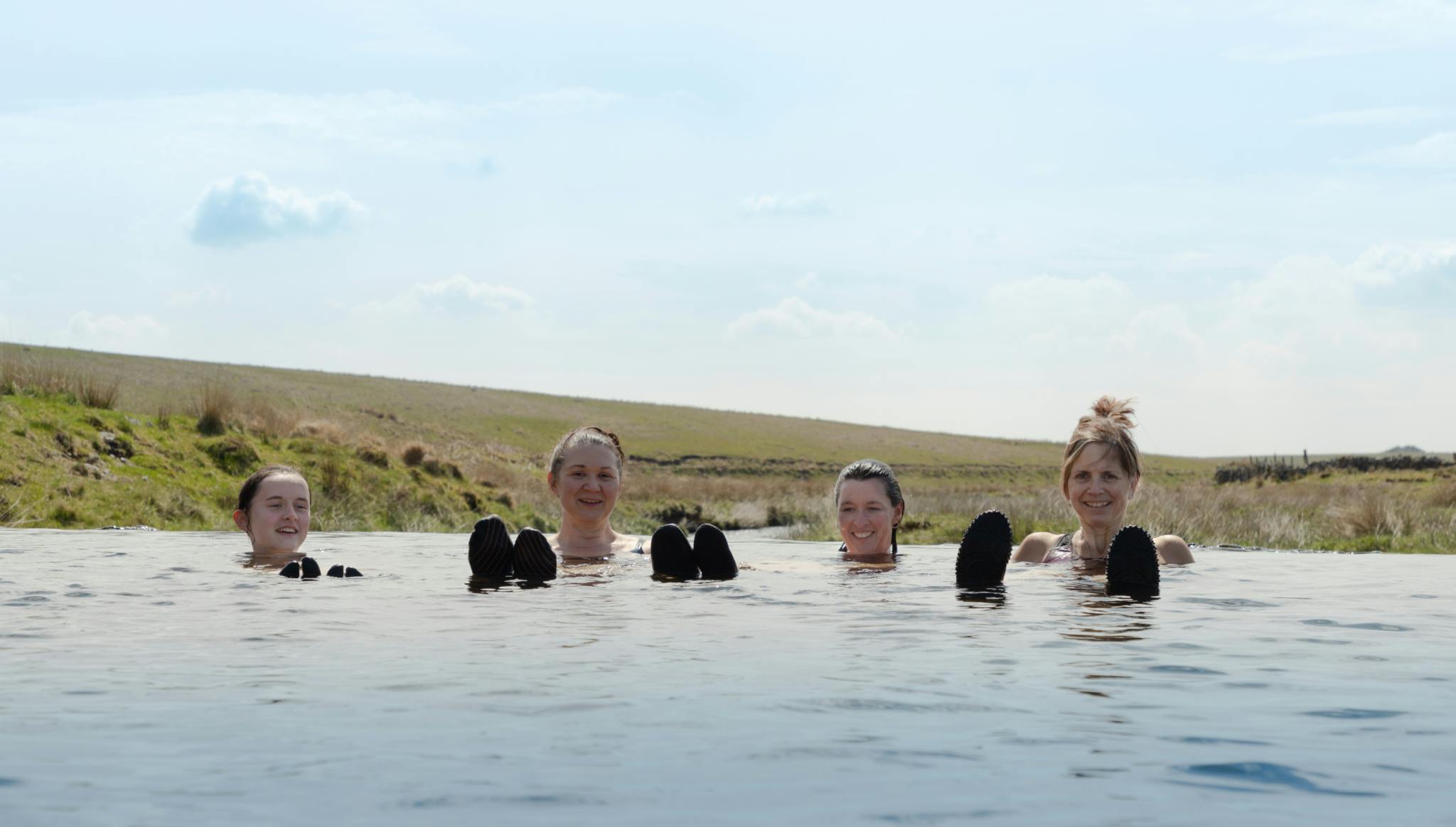 Image shows four women floating in natural water with smiles on their faces and Dartmoor in the background. You can see some of their swimming booties in the foreground, popping through the water.