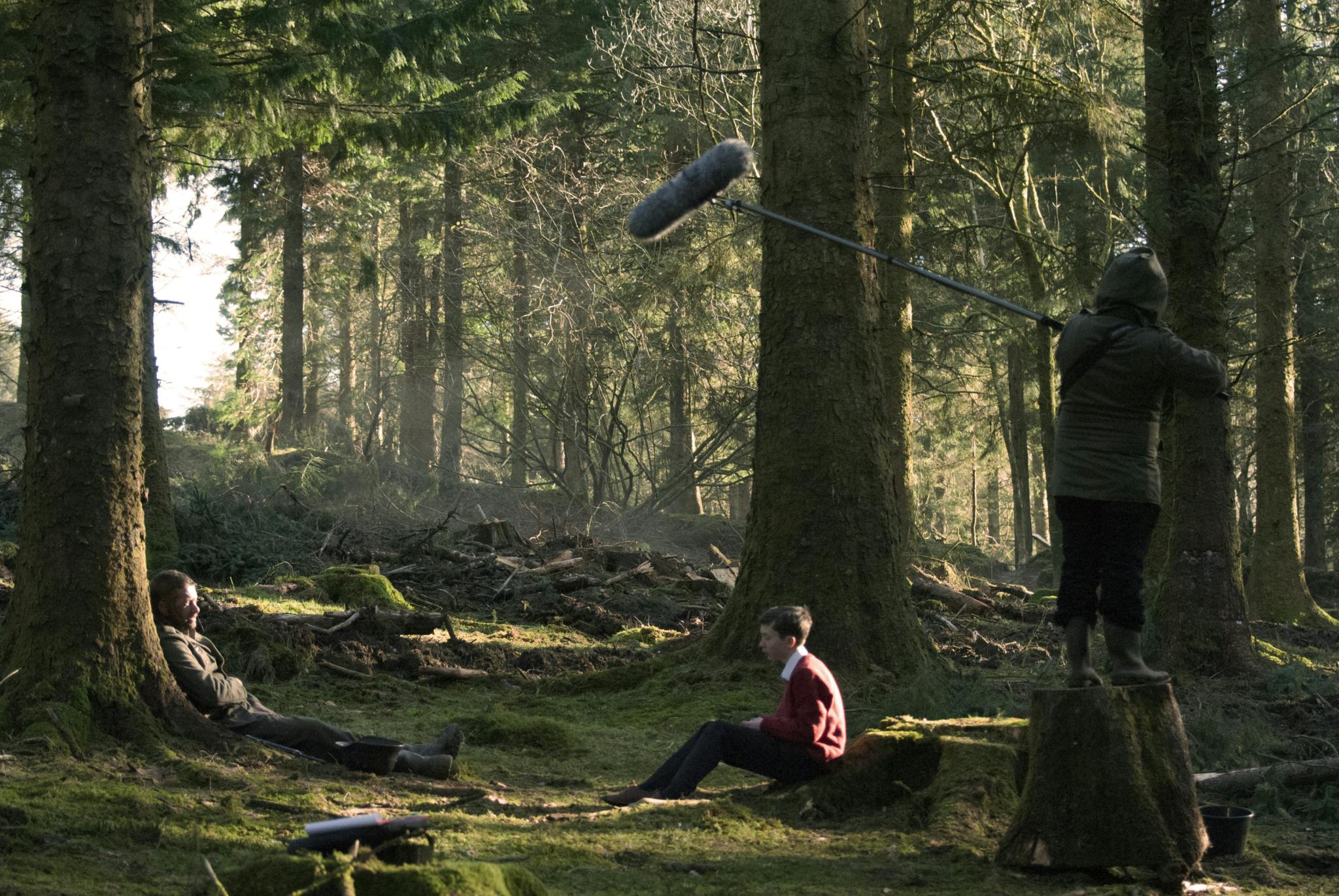A film crew capture a scene with a young boy leaning against a wooden stump on the forest floor, facing a wounded man. Light filters through the tree leaves and a student holding a sound mic dips into shot.