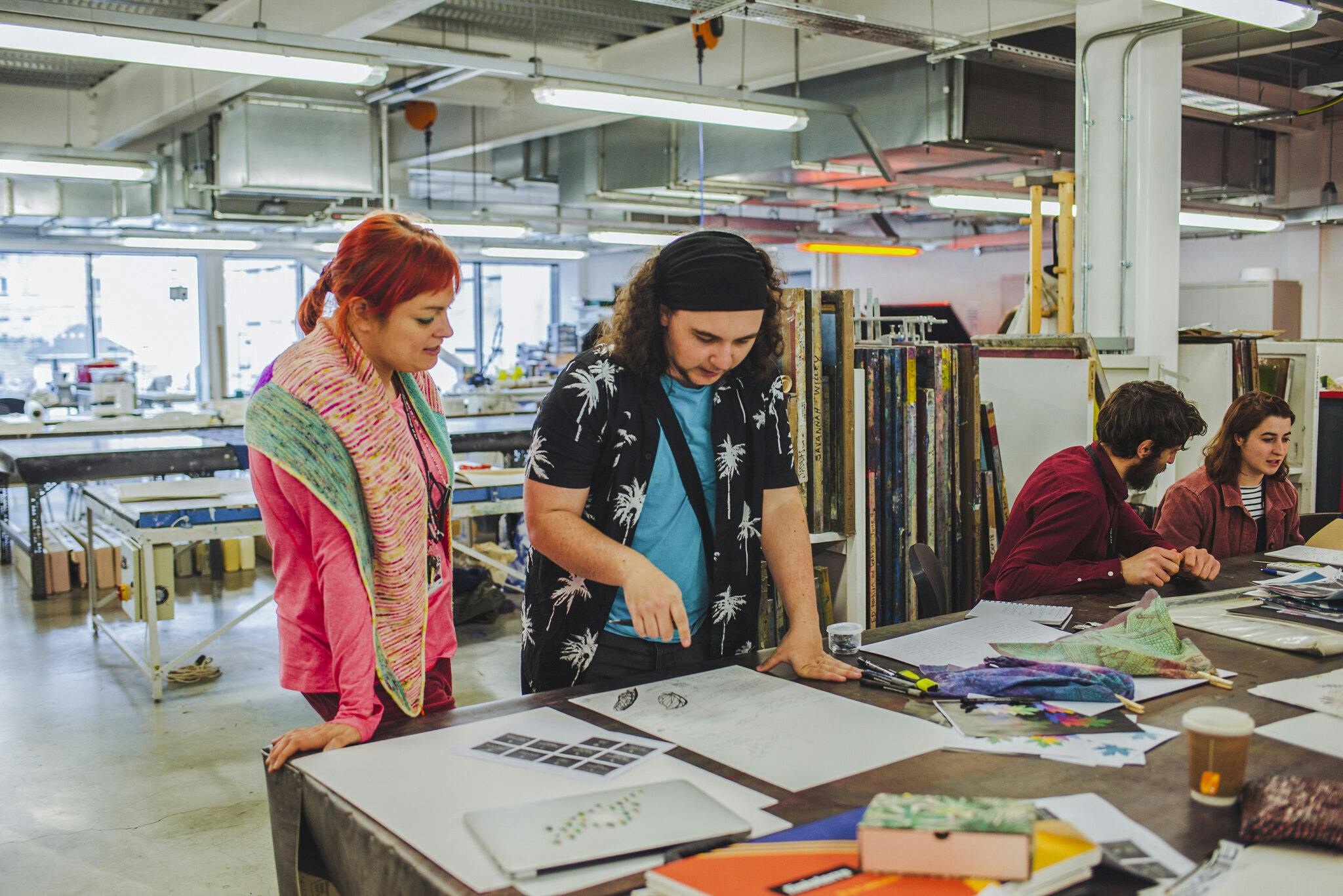 Lecturer Becky Dodman stands next to a textiles student and discuss his work laid out on the print table in front of them, a mix of bright colours and fabrics in our bright and open print studio