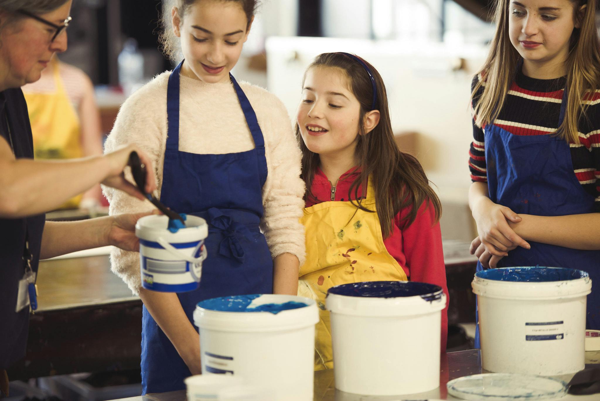 A staff member leans in to show a group of young girls a pot of paint. The girls smile and lean in to see the colour up-close. Three more paint tubs are in the foreground.