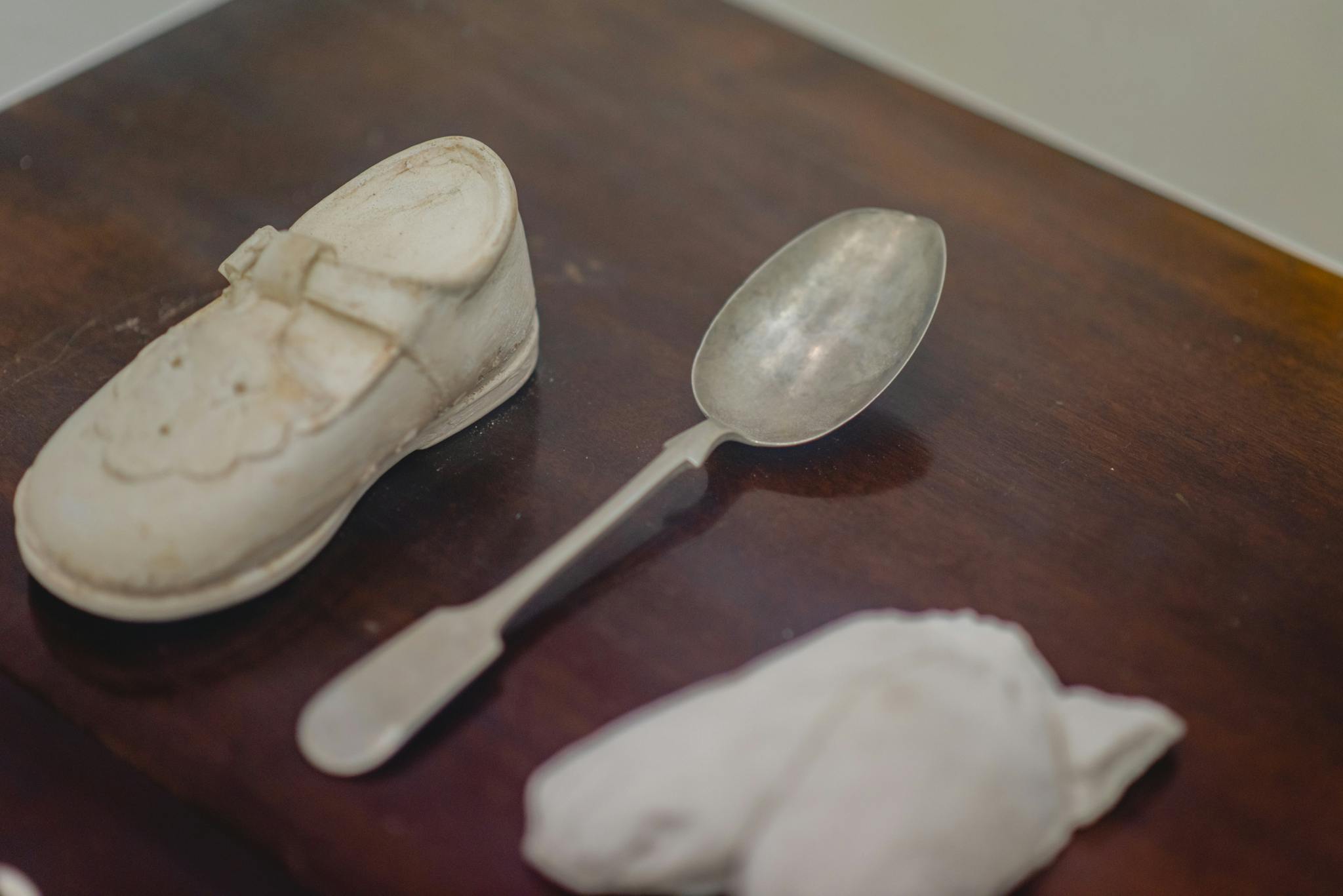 Image shows small objects on a wooden table, a small child's shoe, a spoon and something in the foreground that is too blurry to describe