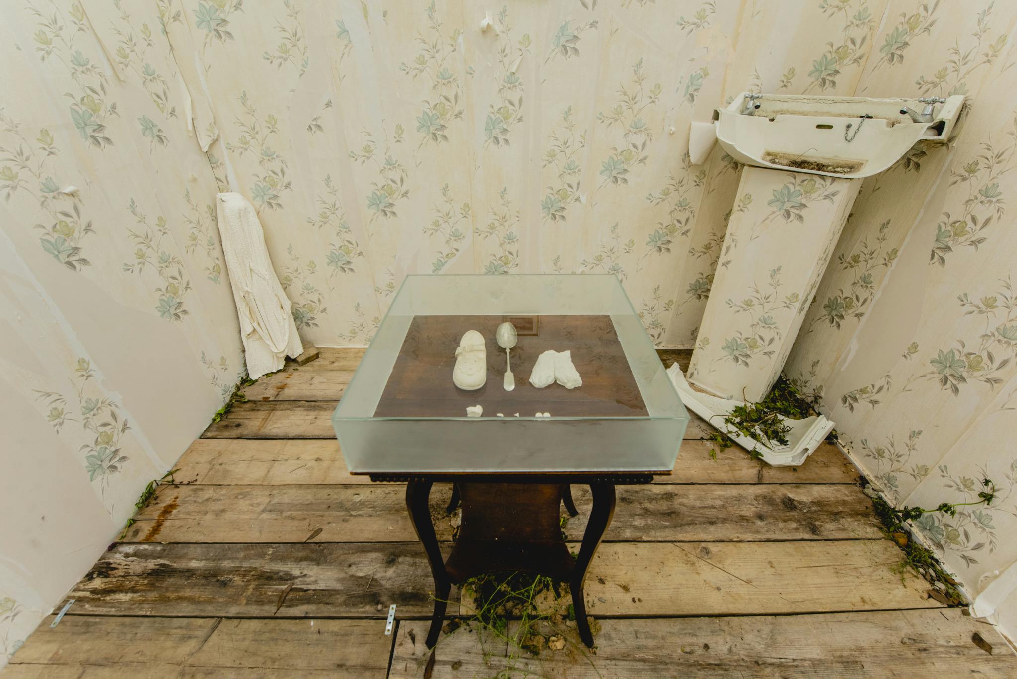 Image shows an art installation with a broken ceramic sink in the background surrounded by peeling wallpaper with a box of sentimental items in a glass box in the foreground