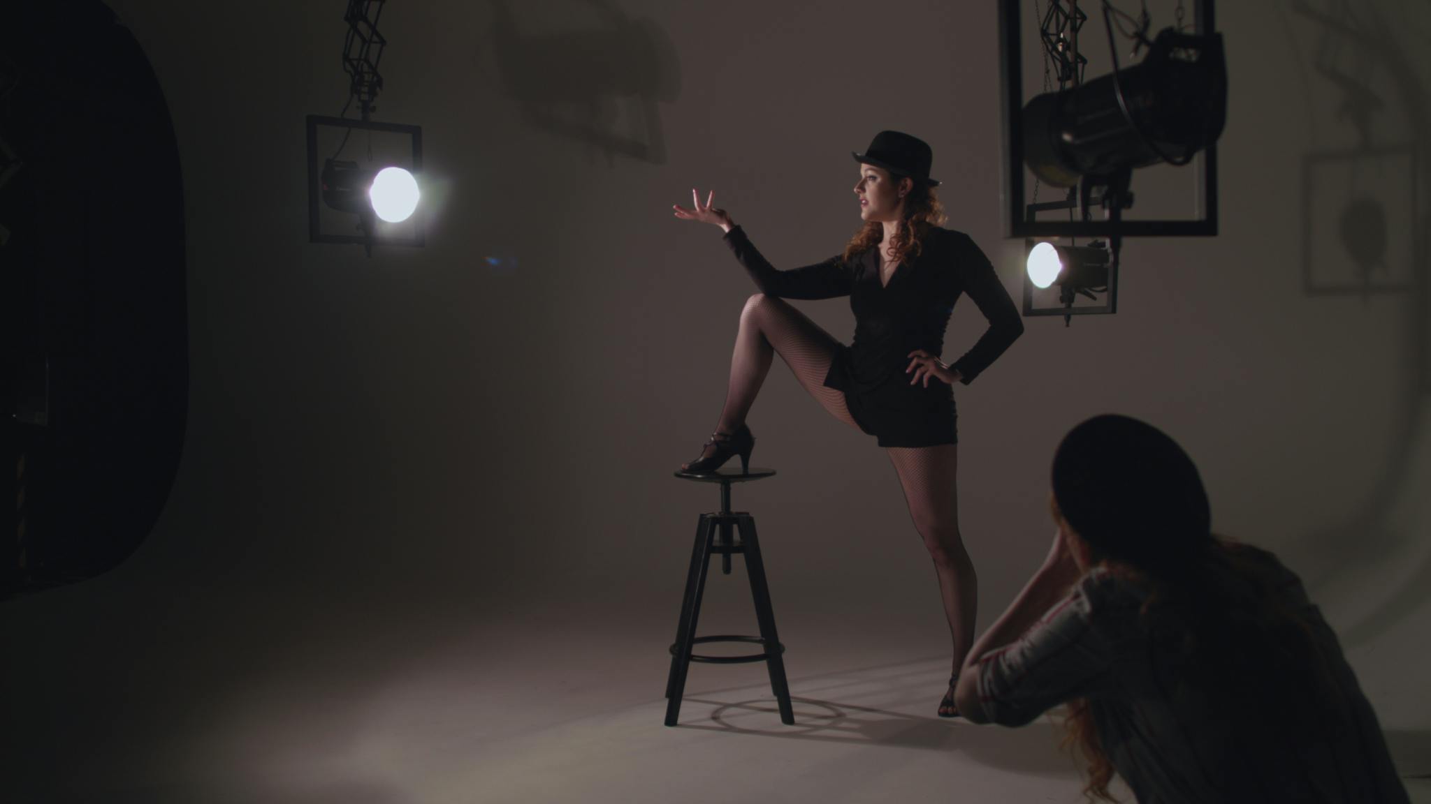 An image featuring a woman in the middle of the shot with her foot up on a stool, wearing a costume, she is surrounded by stage lights and in the foreground a photographer takes her image