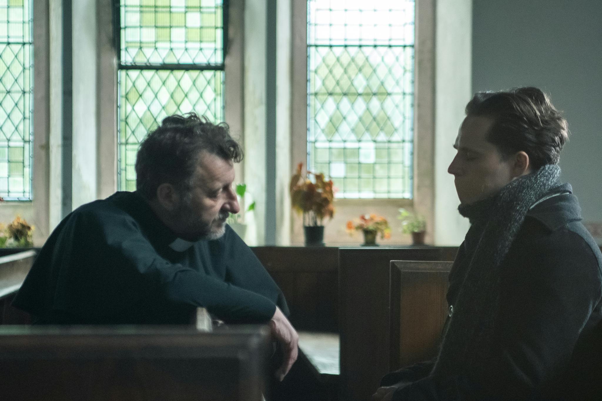 Picture features two characters in a church, on the left is a older man, a priest, speaking to a younger man on the right who's wearing a scarf and a heavy coat