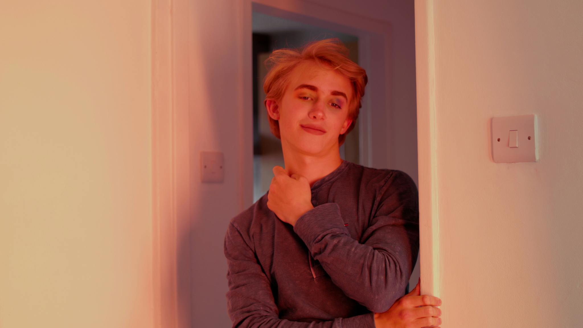 Image shows a young individual with blonde hair in a door frame with soft pink and purple lighting